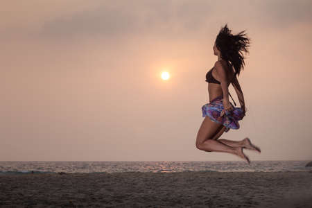 beautiful brunette young girl in style flower skirt and and brown bra jumping on the sand and looking on a sunset on the beachの写真素材