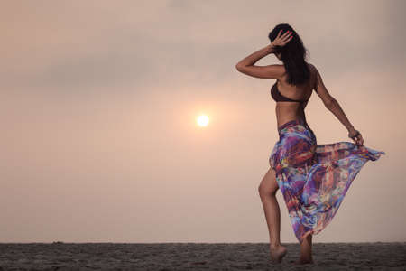 beautiful brunette young girl in style flower skirt and and brown bra standing on the sand and looking on a sunset on the beachの写真素材