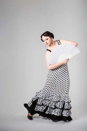 young beautiful brunette female spanish flamenco dancer in black and white flamenco dress dancing with white fan in studio on gray backgroundの写真素材