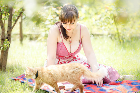 young beautiful brunette girl in pink dress sit on green grass in the garden with red catの写真素材