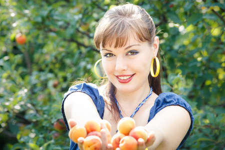 beautiful young brunette girl in blue dress with peach in her hands stand in green garden smile and look at the cameraの写真素材