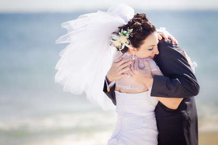 beautiful brunette bride  in white wedding dress hold strong her groom walk on the beach with blue sea on backgroundの写真素材