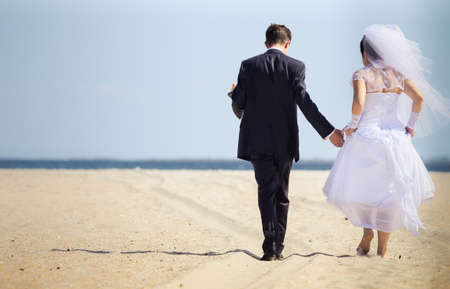 beautiful brunette bride  in white wedding dress and her groom walk on the beach with blue sea on backgroundの写真素材