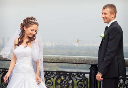 beautiful brunette bride in white wedding dress and young handsome groom in black suit stand on big city backgroundの写真素材
