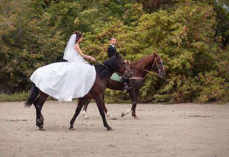 young charming brunette bride in white wedding dress and handsome groom in black suit rides on horsesの写真素材