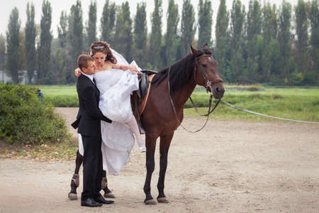 handsome groom in black suit help his young charming brunette bride in white wedding dress get from the horseの写真素材
