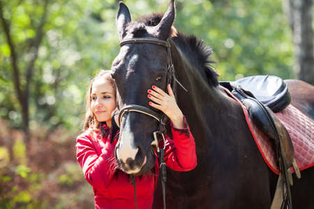 young beautiful brunette girl  in red dress hugg black horse in green forestの写真素材