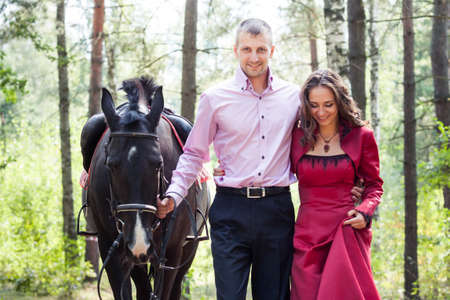 handsome man in pink shirt hold young beautiful brunette girl in long red dress walk with black horse in green forestの写真素材