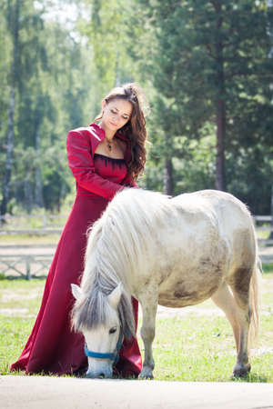 young beautiful brunette girl in long red dress play with white pony horse in green fieldの写真素材