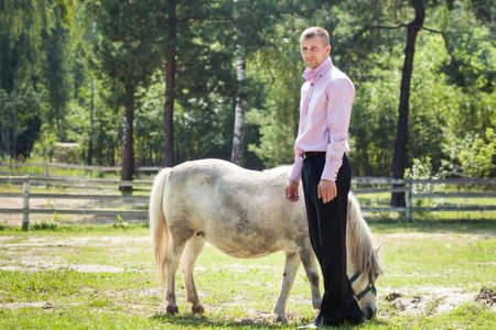 handsome man in pink shirt play with white pony horse in green forestの写真素材