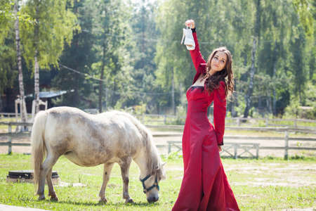 young beautiful brunette girl in long red holding white heels shoes in her hand with with white pony horse in green fieldの写真素材