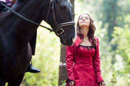 young beautiful brunette girl in red dress and black horse in green forestの写真素材