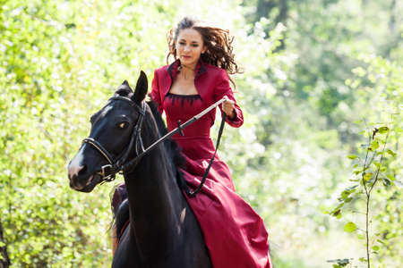 young beautiful brunette girl in red dress ride on the black horse in green forestの写真素材