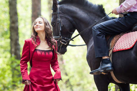 young beautiful brunette girl in red dress walk and handsome man in pink shirt ride on the black horse in green forestの写真素材