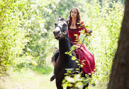 young beautiful brunette girl in red dress ride on the black horse in green forestの写真素材