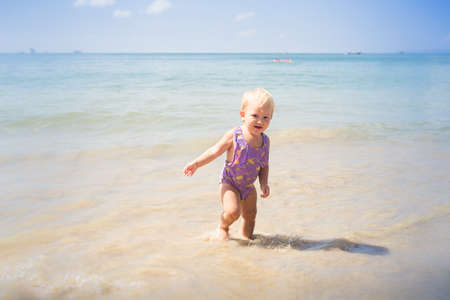 small blonde baby girl in purple swim suit play with waves on seashoreの写真素材