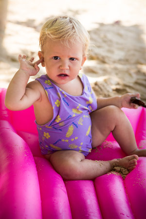 small blonde baby girl in swim suit sit on pink mattress on the beach