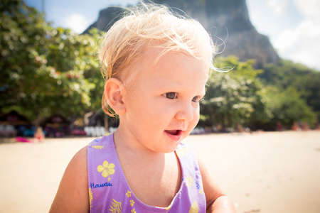 portrait of small blonde baby girl in purple swim suit play on seashore closeupの写真素材