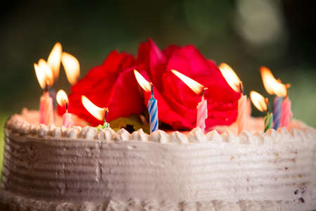 candle burning on a cream white creamy delicious cake decorated with three red roses on the topの写真素材