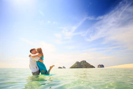 handsome man hold his beautiful blonde wife in elegant dress stand in water with green mountain on backgroundの写真素材