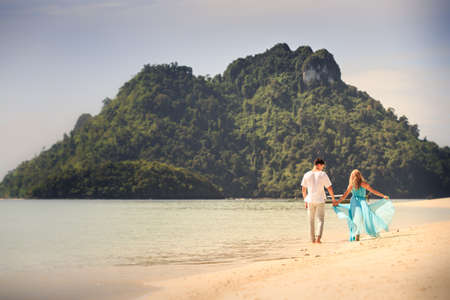 beautiful blonde woman in elegant dress walk with her handsome husband on island beach with green mountain on backgroundの写真素材