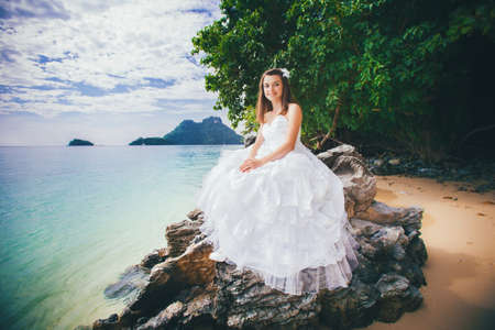 young pretty brunette bride in elegant wedding dress sits on rock against azure sea and islandの写真素材