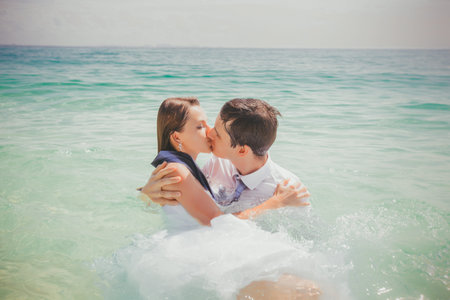 dressed bride and groom kiss completely in shallow water against azure seaの写真素材