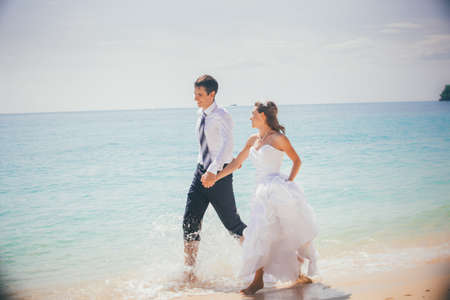 young brunette bride and groom run holding hand in hand on sandy beach in shallow against azure sea and rocky islandの写真素材