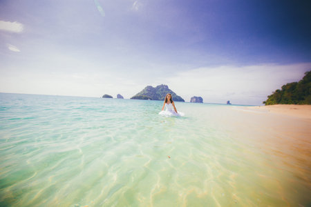 young brunette bride in white dress smile standing in shallow azure water against rocky islandsの写真素材