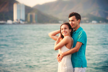 brunette girl in white dress and guy stand and hug standing on large stone at background of azure sea at dawnの写真素材
