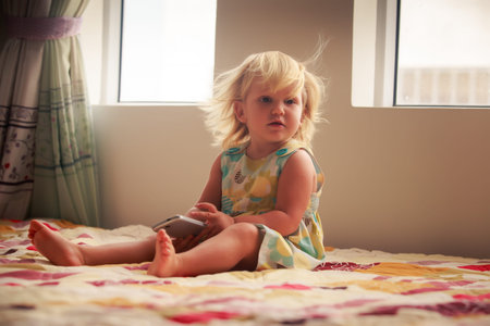 small blonde two years old girl plays with smartphone sitting on couch near windowの写真素材