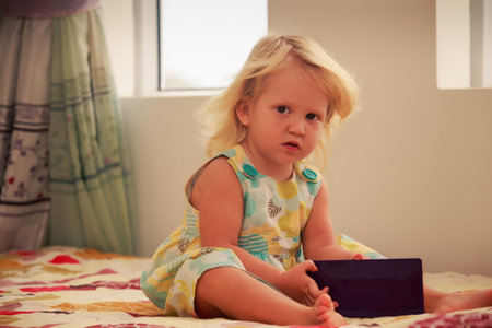 small blonde two years old girl plays with smartphone sitting on couch near windowの写真素材