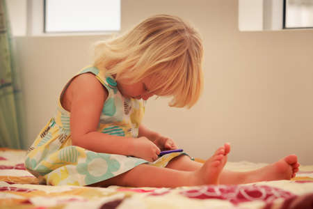 small blonde two years old girl plays with smartphone sitting on couch near windowの写真素材