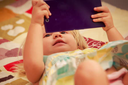 little pretty blonde two years old girl plays with smartphone lay on colored couchの写真素材