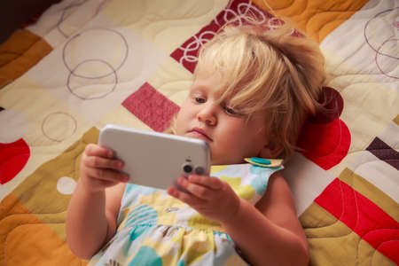 pretty blonde two years old girl plays with smartphone lay on colored couchの写真素材