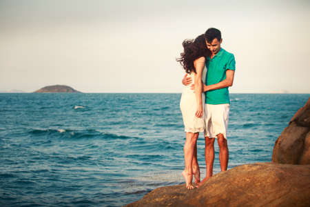 brunette girl in white dress and guy stand and hug standing on large stone at background of azure sea at dawnの写真素材