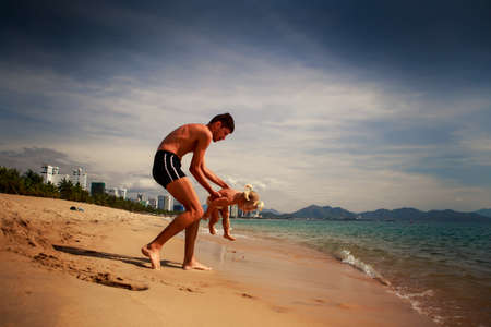 european young father plays with little daughter in shallow water on sand beach against modern resort cityの写真素材
