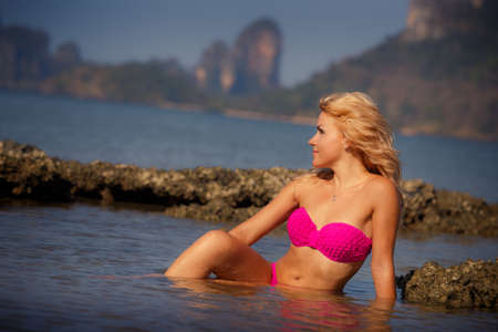 blonde longhaired girl in swimming suit half in sea water at stones against cliffy islandの写真素材