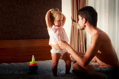 father plays with and speaks to little blonde daughter in pink dress on sofa near toys against windowの写真素材