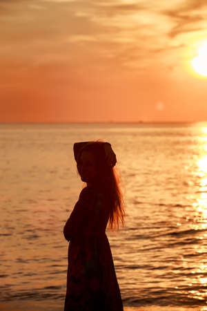 slim longhaired girl in long dress side view on beach at background of rising over sea sun and cloudsの写真素材