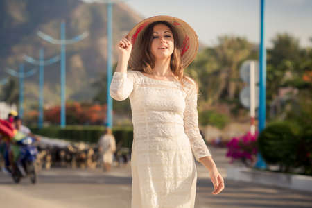 slim blonde girl in Vietnamese national white long dress and hat walks along road against flock of goatsの写真素材
