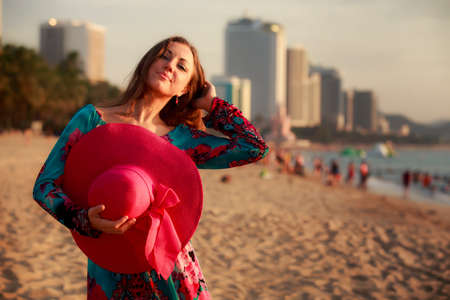 slim brunette longhaired girl in colorful dress holds big red hat behind back on beach against city palms and seaの写真素材