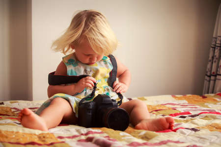 little blonde girl in colorful dress tries to put on photo camera sitting on sofa against white wallの写真素材