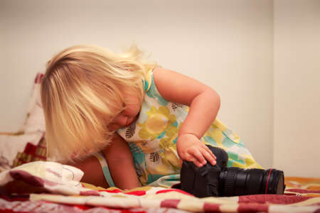 little blonde girl in colorful dress examines  with interest photo camera sitting on sofa against white wallの写真素材