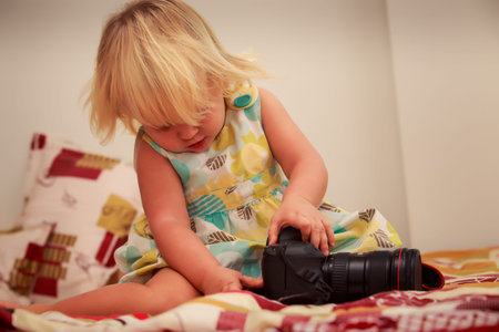 little blonde girl in colorful dress examines  with interest photo camera sitting on sofa against...