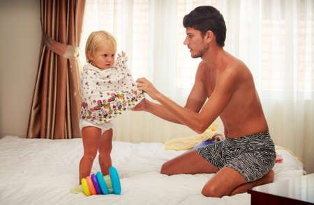 father puts on new colourful dress to little blonde daughter on sofa against bright windowの写真素材