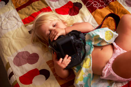 little blonde girl in colorful dress lies on sofa and examines with interest photo cameraの写真素材