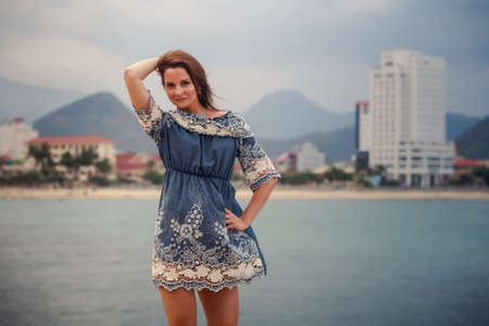 young brunette long-legged girl in short frock with lace barefoot stands tiptoe on stone against sea and resort cityの写真素材