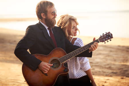 guitarist in black suit and blonde girl in purple dress standing on sandy beach against treeの写真素材