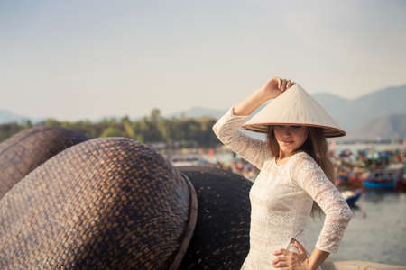 slim blonde girl in Vietnamese national white long dress touches hat by barrier of embankment against boats mountainsの写真素材
